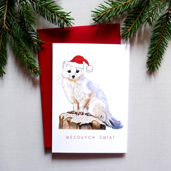Kartka Świąteczna z uroczym Zwierzakiem, Biały Gronostaj w mikołajkowej czapeczce, wraz z czerwoną kopertą na tle świątecznych dekoracji.